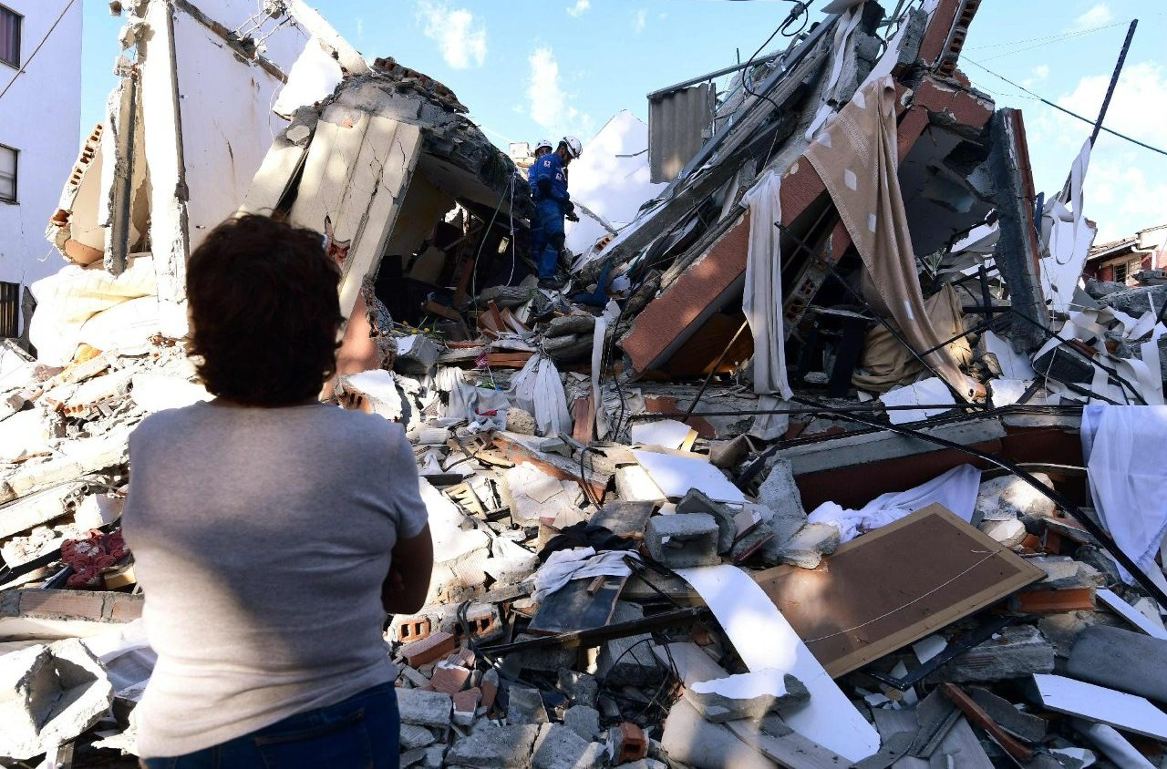 A person looking toward a collapsed building and rubble, with a rescue worker among the debris